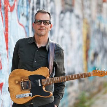 Picture of Luca Benedetti in front of a wall with graffiti. He is wearing glasses and holding a guitar and looking at the camera.