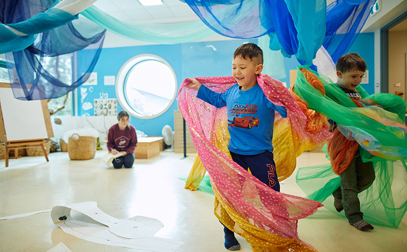 Two children playing with cloth ribbons while an ECCE student observes.